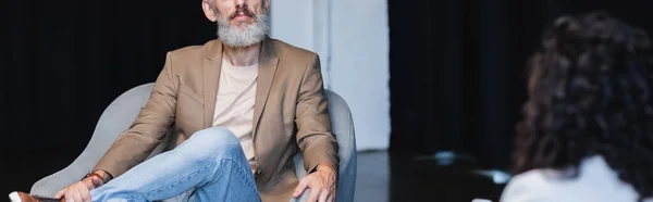 Vista recortada de empresario confiado sentado en sillón cerca borrosa periodista afroamericano durante la entrevista, pancarta - foto de stock