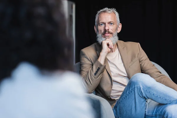 Selbstbewusster Geschäftsmann sitzt im Sessel neben verschwommenem afrikanisch-amerikanischem Journalisten beim Interview — Stockfoto