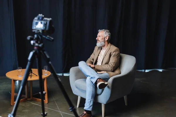 Homme d'affaires confiant assis dans un fauteuil près de l'appareil photo numérique flou sur trépied — Photo de stock