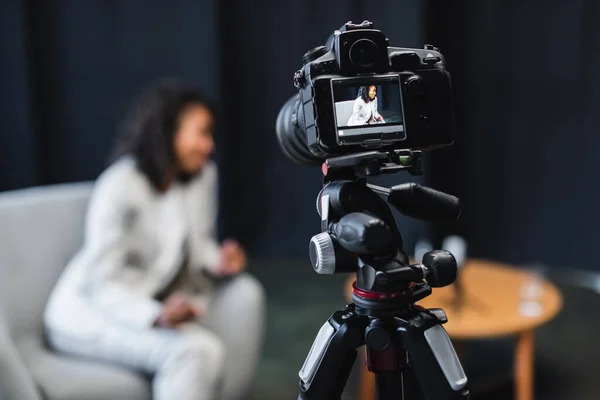 Modern digital camera on tripod with african american journalist sitting in armchair on screen — Stock Photo