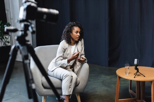 Periodista afroamericano sentado en sillón y hablando cerca de cámara digital borrosa en trípode - foto de stock