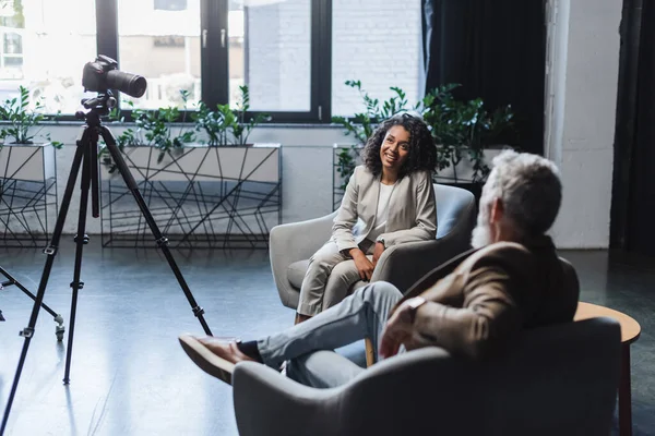 Glücklich afrikanisch-amerikanischer Journalist und unscharfer Geschäftsmann im Gespräch während eines Interviews in der Nähe der Digitalkamera auf Stativ — Stockfoto