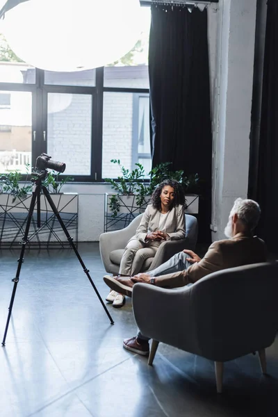 Lockige afrikanisch-amerikanische Journalistin schaut Geschäftsmann beim Gespräch in der Nähe von Digitalkamera auf Stativ an — Stockfoto