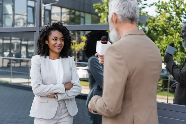 Verschwommene und bärtige Reporterin mit Mikrofon in der Nähe lächelnder afrikanisch-amerikanischer Geschäftsfrau — Stockfoto