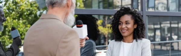 Расплывчатый и бородатый репортер с микрофоном, говорящий рядом с улыбающейся африканской бизнесвумен, баннер — стоковое фото