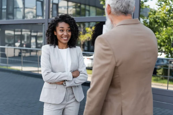 Lächelnde afrikanisch-amerikanische Geschäftsfrau mit verschränkten Armen neben verschwommenem Reporter — Stockfoto