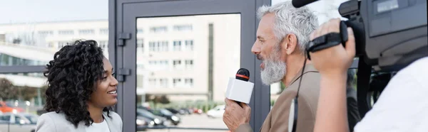 Reporter mit Mikrofon interviewt glückliche afrikanisch-amerikanische Geschäftsfrau in der Nähe verschwommener Kameramann mit Videokamera, Banner — Stockfoto