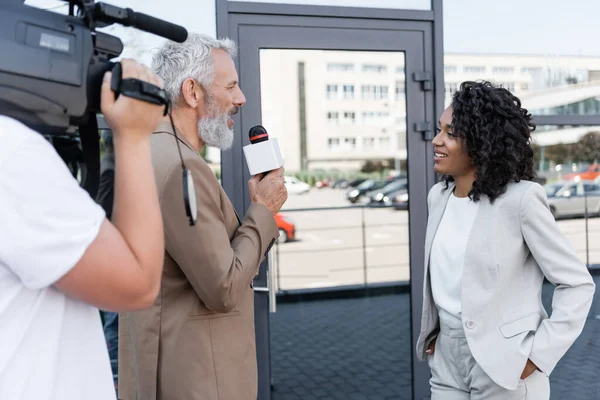 Репортер з інтерв'ю з мікрофоном щаслива афроамериканська бізнес-леді біля розмитого оператора з відеокамерою — стокове фото
