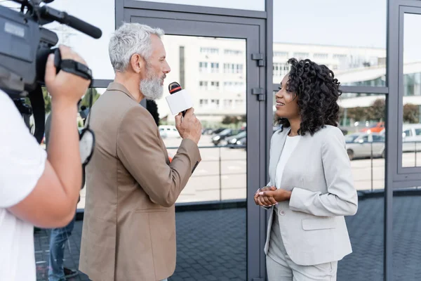 Giornalista con microfono intervistando sorridente donna d'affari afroamericana vicino cameraman offuscata con videocamera — Foto stock