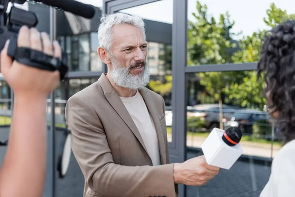 Bearded reporter with microphone interviewing african american businesswoman near blurred cameraman with video camera — Stock Photo