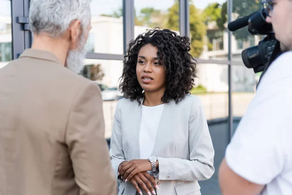 Reporter interviewt hübsche afrikanisch-amerikanische Geschäftsfrau in der Nähe verschwommener Kameramann mit Videokamera — Stockfoto