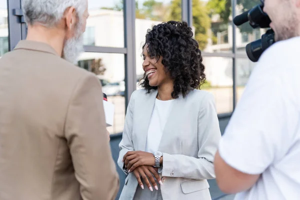 Reporter interviewing cheerful african american businesswoman near blurred cameraman with video camera — Stock Photo