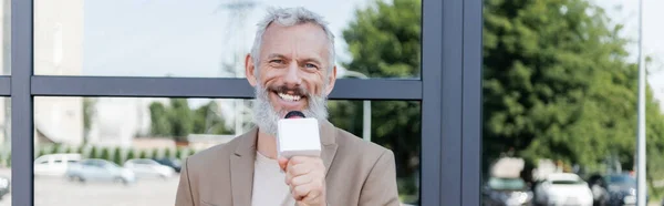 Glücklicher Reporter im Blazer hält Mikrofon in der Hand und macht Reportagen in der Nähe des Gebäudes, Banner — Stockfoto