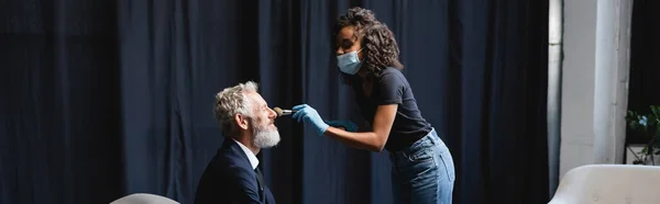 Maquilleur afro-américain bouclé en masque médical et gants en latex appliquant de la poudre visage sur le visage de l'homme d'affaires, bannière — Photo de stock