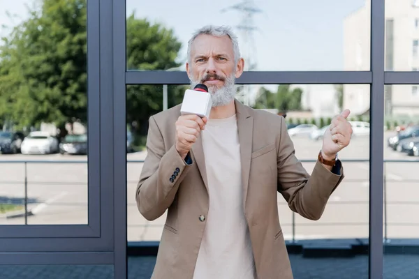 Бородатый репортер в блейзере держит микрофон и указывает пальцем на здание — стоковое фото
