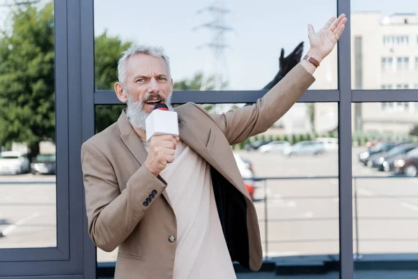 Bearded reporter in blazer holding microphone and pointing with hand at building — Stock Photo