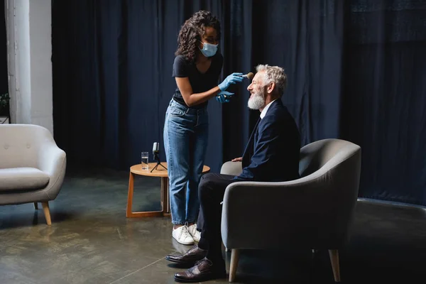 Maquilleur afro-américain en masque médical appliquant de la poudre visage sur homme d'affaires joyeux en studio — Photo de stock