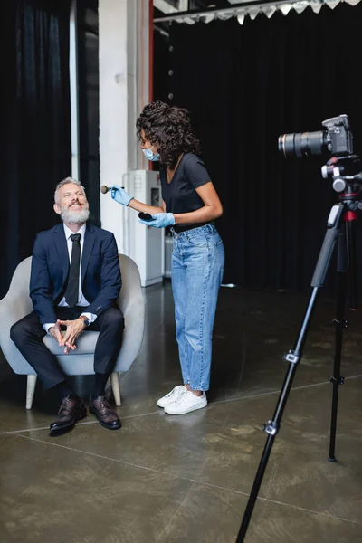 Artista de maquiagem afro-americana em máscara médica aplicando pó facial no rosto de empresário sorridente em estúdio — Fotografia de Stock