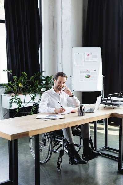 Empresario discapacitado positivo escribir en el cuaderno durante la conversación en el teléfono inteligente en la oficina - foto de stock