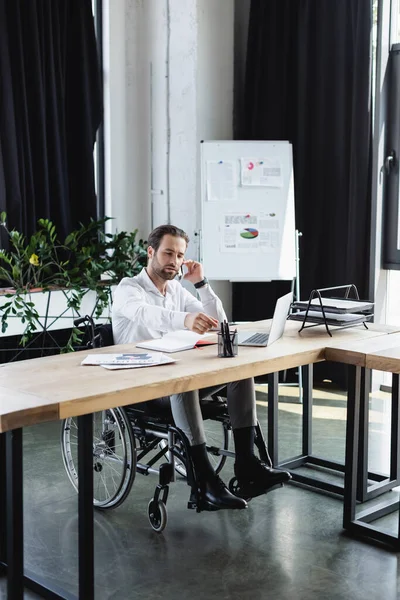 Behinderter Geschäftsmann greift zum Stift beim Telefonieren in der Nähe von Laptop im Büro — Stockfoto