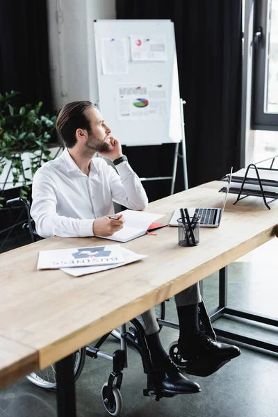 Thoughtful disabled businessman looking away near laptop and blurred flip chart in office — Stock Photo