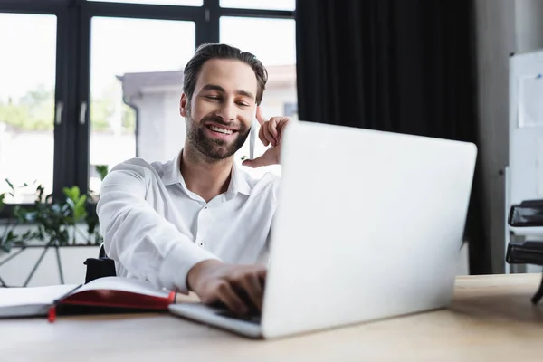 Happy businessman using blurred laptop while talking on smartphone in office — Stock Photo