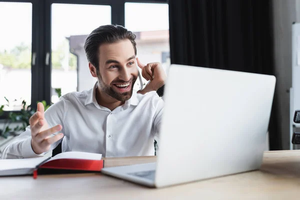 Homem de negócios feliz gesticulando enquanto fala no smartphone perto do laptop desfocado no escritório — Fotografia de Stock
