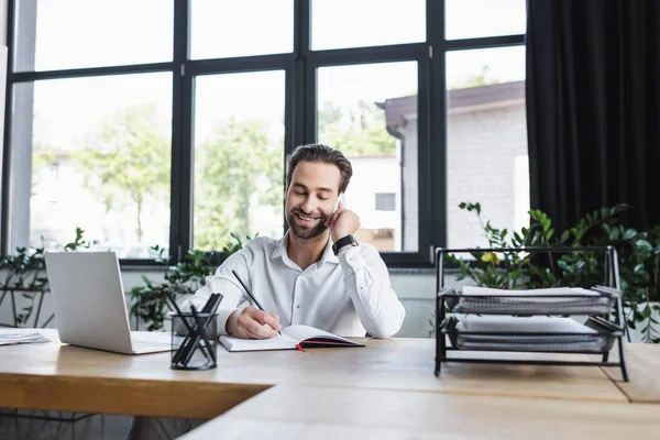 Uomo d'affari sorridente che scrive nel notebook mentre parla sul telefono cellulare vicino al computer portatile — Foto stock