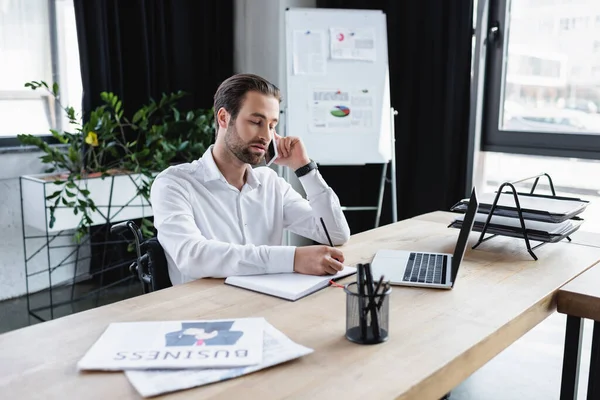 Handicapped businessman talking on mobile phone near laptop and newspapers on desk — Stock Photo