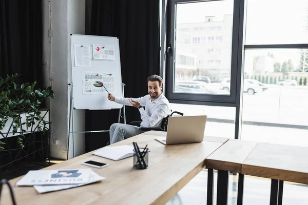 Homme d'affaires handicapé souriant pointant vers l'infographie sur tableau à feuilles mobiles pendant l'appel vidéo sur ordinateur portable — Photo de stock