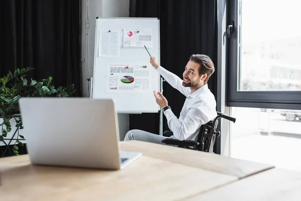 Homme d'affaires handicapé souriant tout en pointant vers flip chart lors de la vidéoconférence sur ordinateur portable flou — Photo de stock