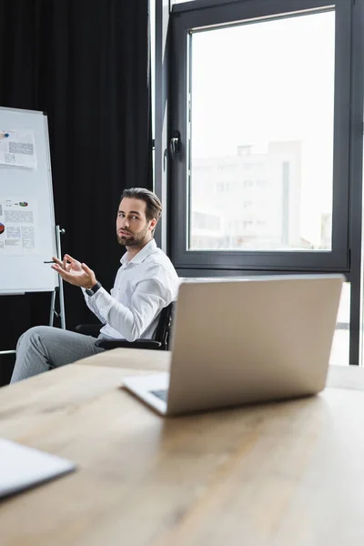 Jeune homme d'affaires sérieux regardant ordinateur portable flou pendant l'appel vidéo et pointant vers flip chart — Photo de stock