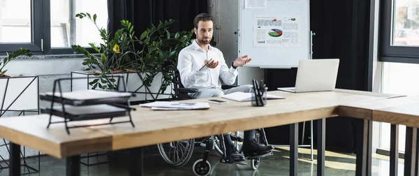 Молодий інвалідний бізнесмен вказує руками під час відеоконференції на ноутбуці біля фліп-чарту з інфографікою, банер — стокове фото