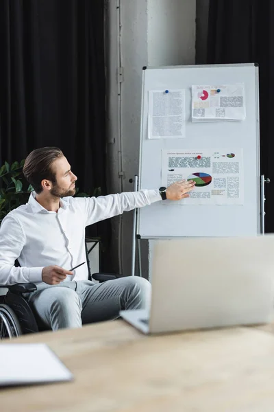 Young disabled businessman pointing at infographics on flip chart during video conference on blurred laptop — Stock Photo