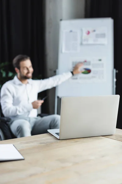 Selective focus of laptop near blurred disabled businessman pointing at flip chart during video call — Stock Photo