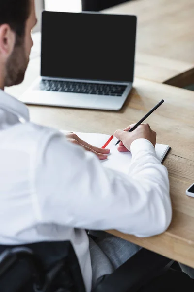 Vista cortada de empresário desfocado escrevendo em notebook perto de laptop com tela em branco — Fotografia de Stock