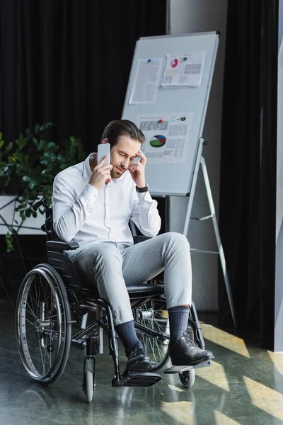 Vue complète de l'homme d'affaires handicapé en fauteuil roulant parlant sur smartphone près tableau à feuilles floues — Photo de stock