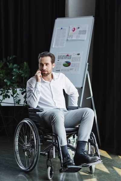 Ganzkörperansicht eines behinderten Geschäftsmannes im Rollstuhl, der im Büro mit dem Smartphone spricht — Stockfoto