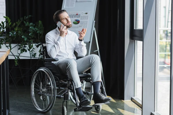 Ganzkörperansicht eines behinderten Geschäftsmannes, der in der Nähe eines Fensters mit einem Handy spricht und ein unscharfes Flipchart zeigt — Stockfoto