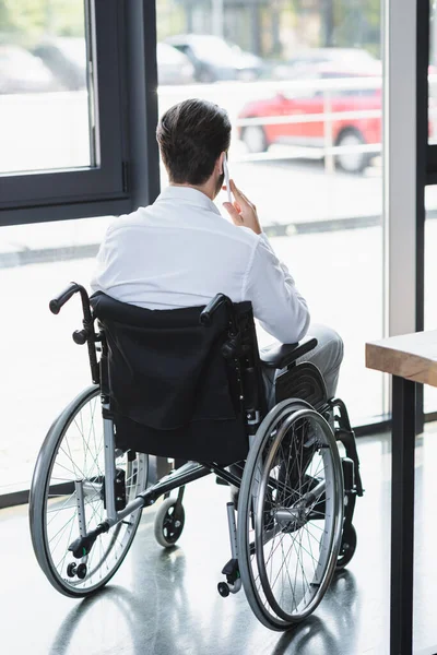 Назад вид инвалида бизнесмен в инвалидной коляске разговаривает на смартфоне возле окна в офисе — стоковое фото