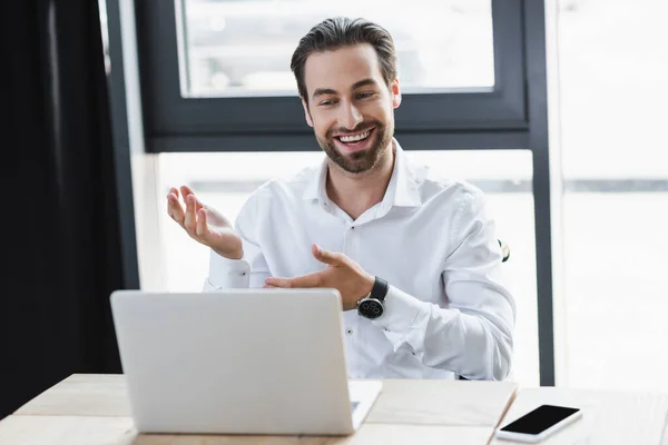 Homme d'affaires positif pointant avec les mains près de l'ordinateur portable et smartphone avec écran blanc dans le bureau — Photo de stock