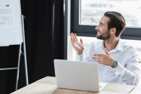 Homem de negócios sorrindo apontando para gráfico flip borrado durante chamada de vídeo no laptop no escritório — Fotografia de Stock