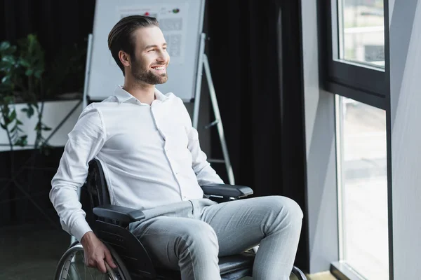 Jovem empresário deficiente em cadeira de rodas olhando para longe e sorrindo no escritório — Fotografia de Stock