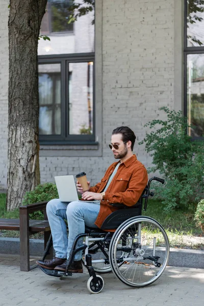 Hombre discapacitado en gafas de sol escribiendo en el ordenador portátil en silla de ruedas al aire libre - foto de stock