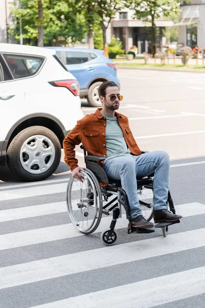Hombre discapacitado en silla de ruedas cruzando la carretera cerca de coches en la ciudad - foto de stock