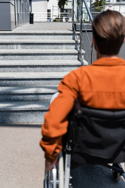 Vista trasera del hombre discapacitado desenfocado en silla de ruedas cerca de escaleras en la calle - foto de stock