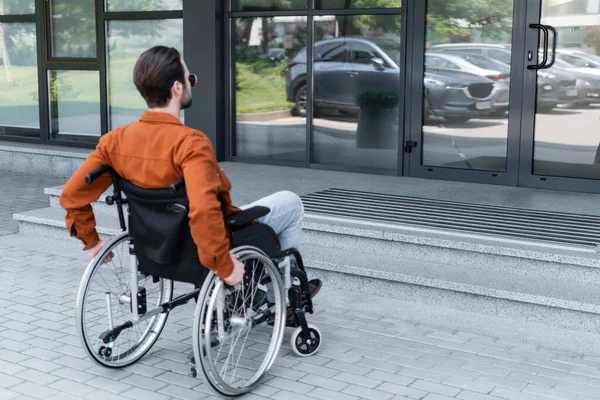 Jovem deficiente em cadeira de rodas perto da entrada do edifício com escadas — Stock Photo