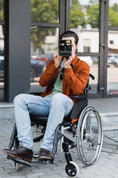 Visão de comprimento total do homem com deficiência em cadeira de rodas tirar foto na câmera vintage na rua — Fotografia de Stock