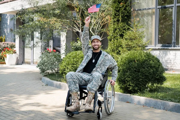 Joven y feliz veterano discapacitado sentado en silla de ruedas con pequeña bandera de EE.UU. en la mano levantada - foto de stock