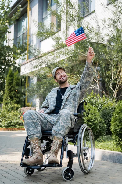 Happy disabled military man in wheelchair holding small usa flag in raised hand outdoors — Stock Photo